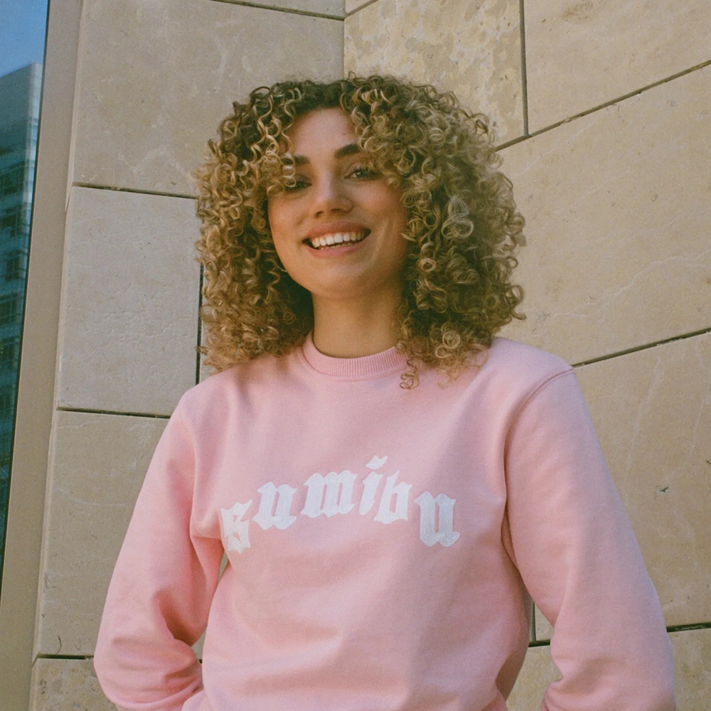 Smiling woman with voluminous blonde curls wearing a light pink sweatshirt with white "gumibu" text.