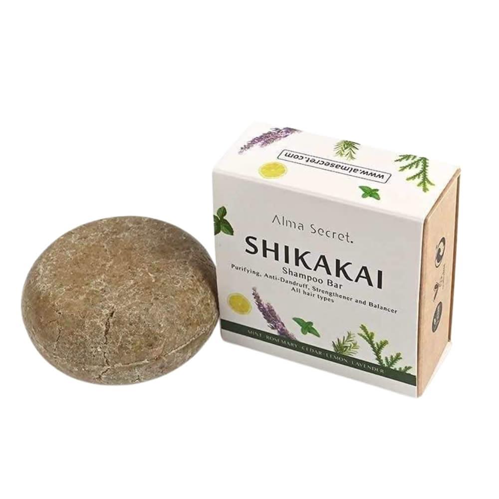 Alma Secret Shikakai solid shampoo bar, round and brown, next to its white box with green botanical design.