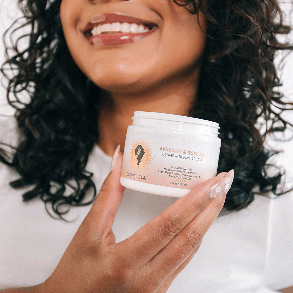 Woman with curly hair holding a white jar of Bounce Curl Avocado & Rose Oil Clump & Define Cream.