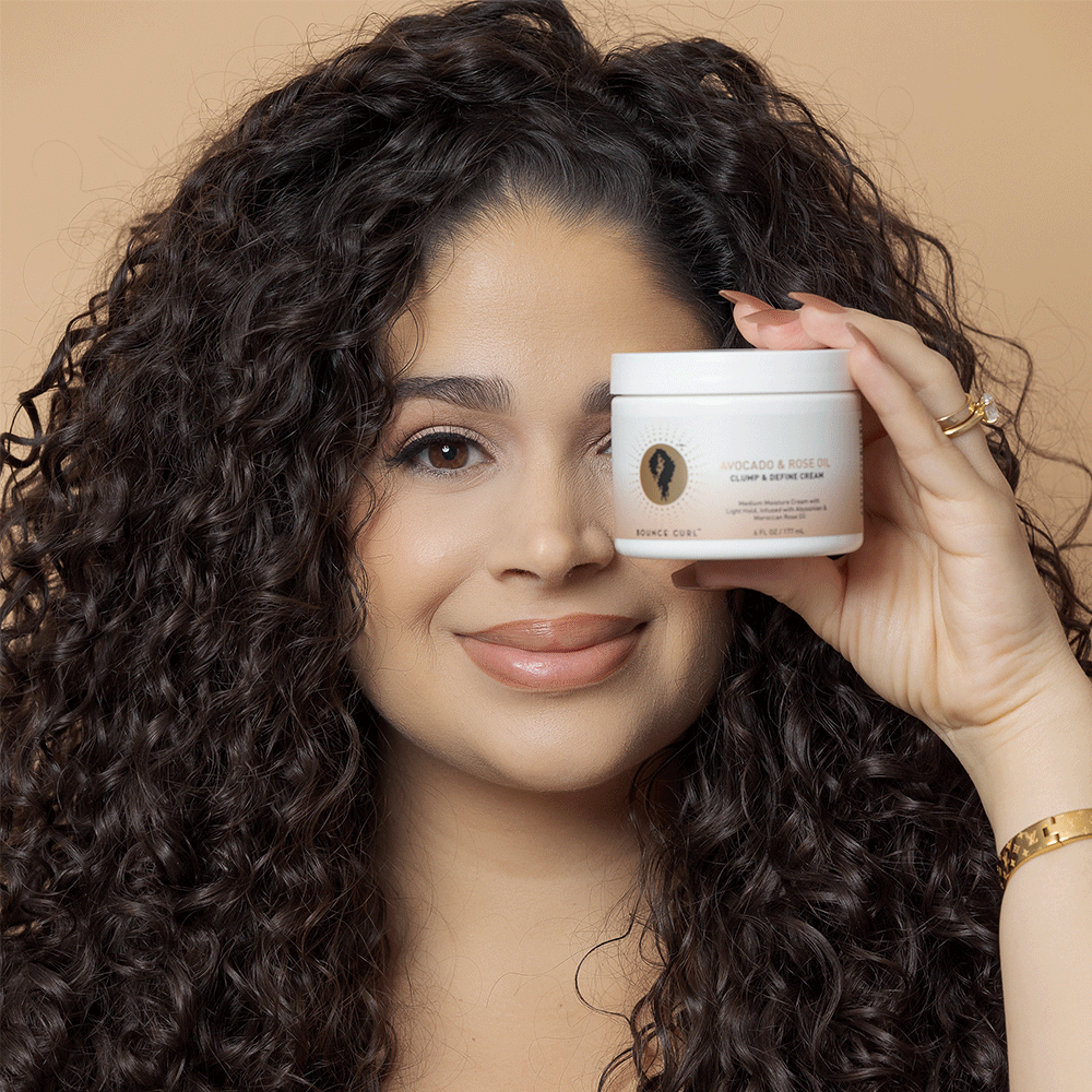 Woman with curly hair holding a white jar of Bounce Curl Avocado & Rose Oil Clump & Define Cream.