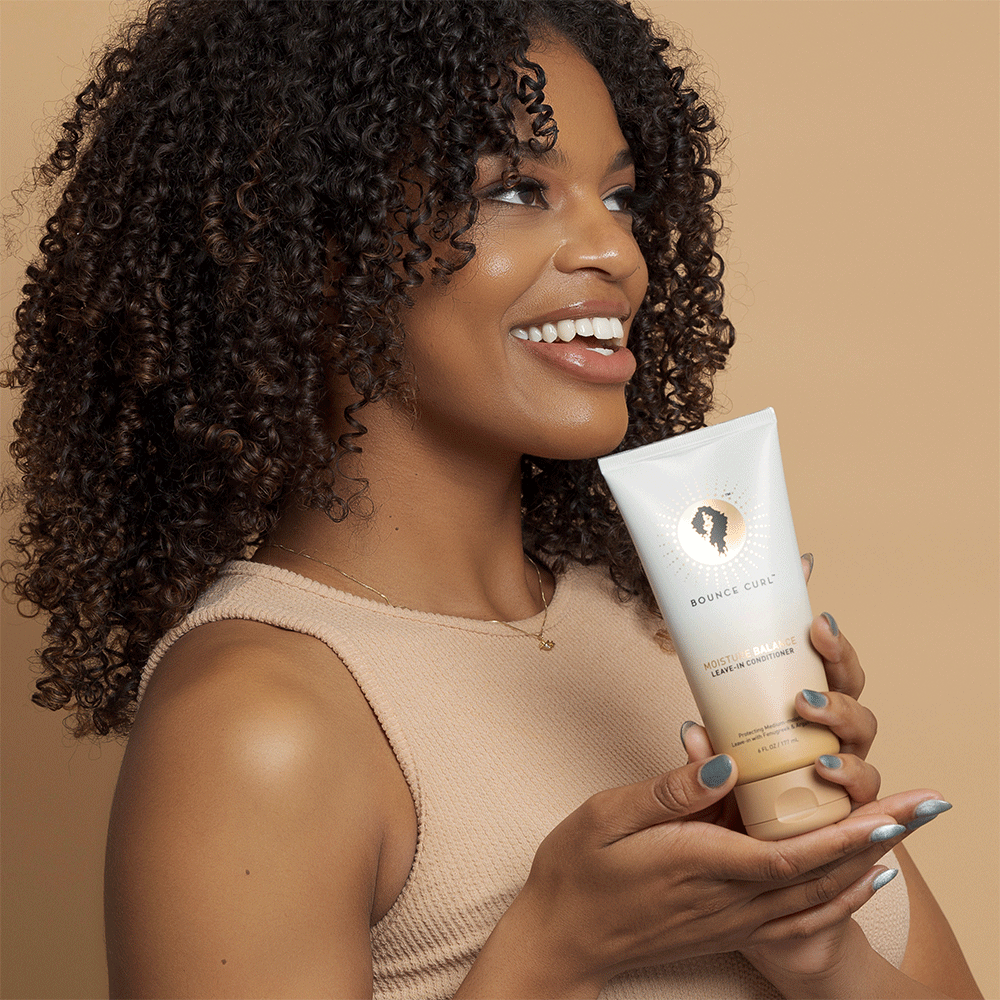 Smiling woman with curly hair holding a white Bounce Curl Moisture Balance Leave-In Conditioner tube.
