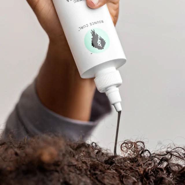 Hand squeezing white Bounce Curl Turmeric Purifying Hair & Scalp Mask bottle onto dark, curly hair.