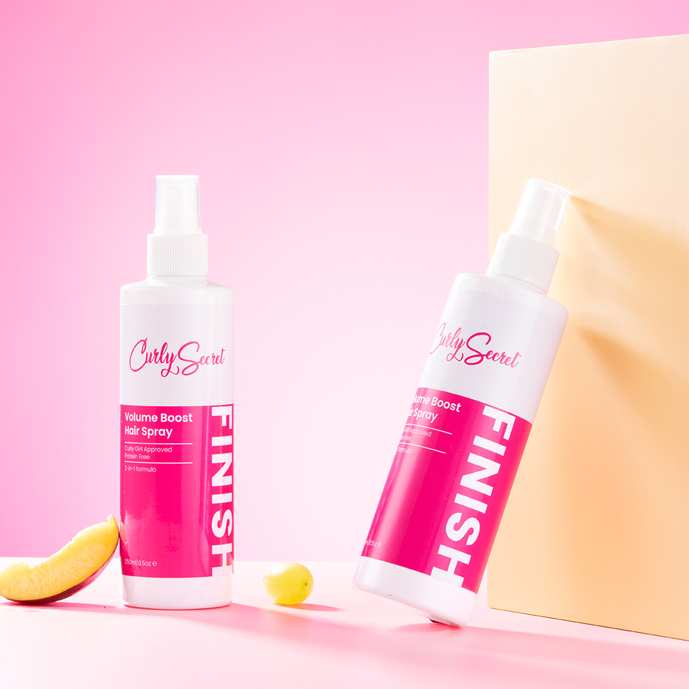Two white Curly Secret Volume Boost Hair Spray bottles with pink labels, one upright, one leaning, on a pink and beige background.