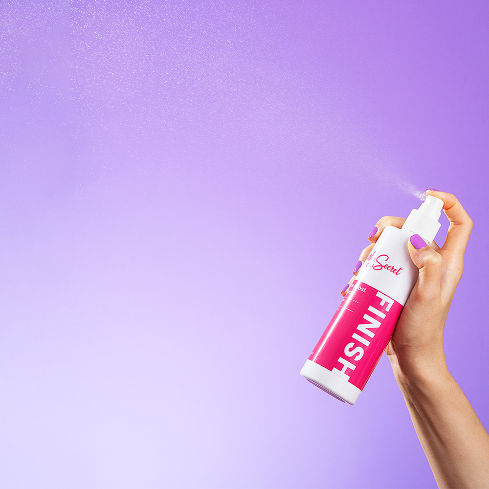 Hand holding a white and pink Curly Secret Volume Boost Hair Spray bottle, spraying mist against a purple background.