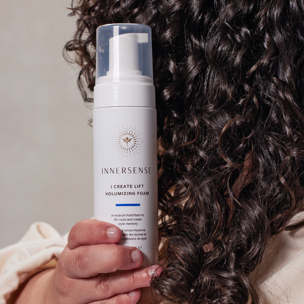 White Innersense I Create Lift Volumizing Foam bottle held against a person's curly brown hair.