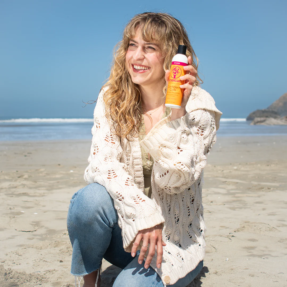 Smiling woman on a beach holding an orange and white Jessicurl Confident Coils bottle.