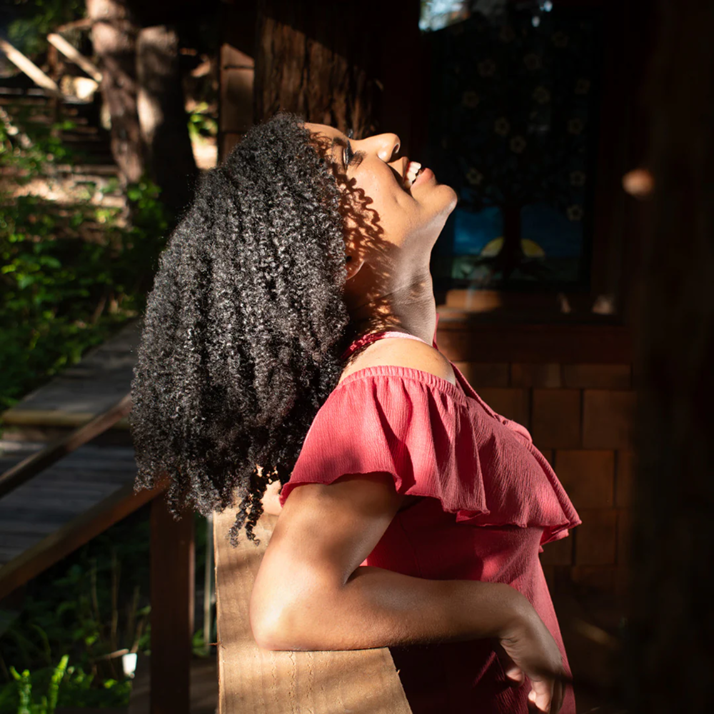 Woman with defined, frizz-free curls in a red off-shoulder top, smiling with her head tilted back in sunlight.