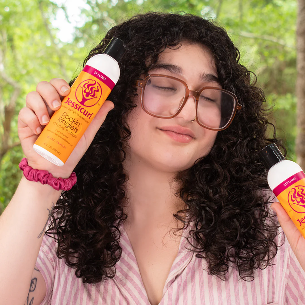 Smiling person with curly hair holding two orange Jessicurl Rockin' Ringlets Styling Potion bottles.