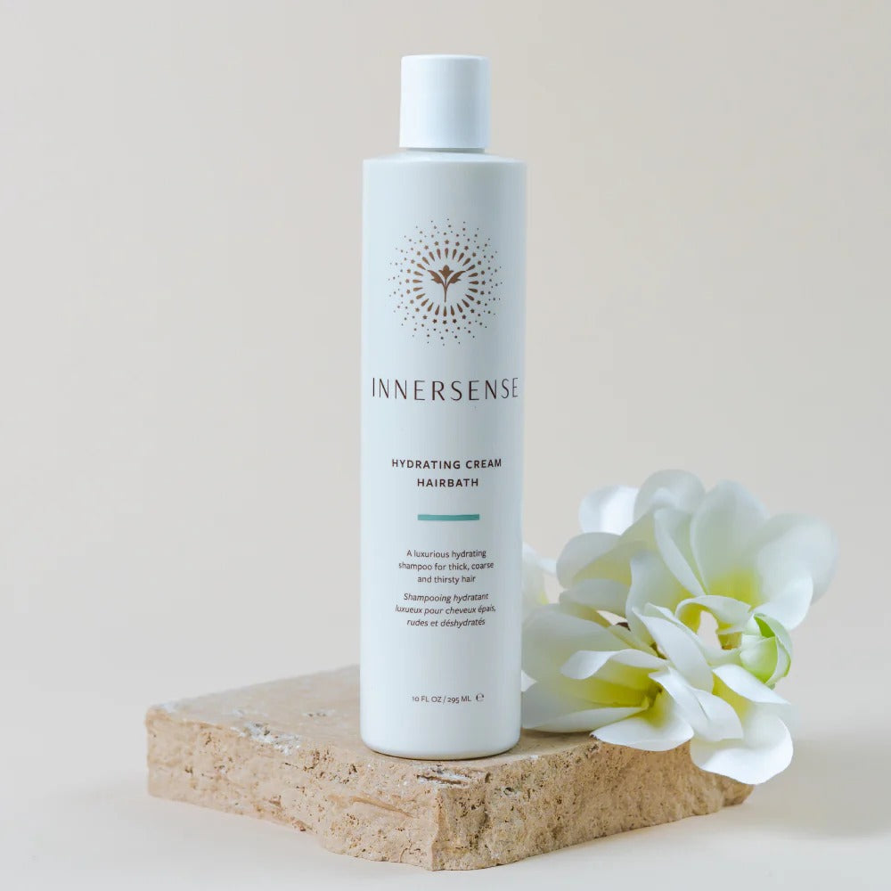 White Innersense Hydrating Cream Hairbath bottle with gold logo, on a beige stone block with white flowers.